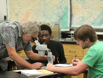 Faculty member looking through a microscope  at rock samples with two male students observing in a classroom setting