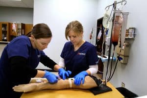 A medical assistant student practicing inserting an IV into a dummy arm while a instructor observes