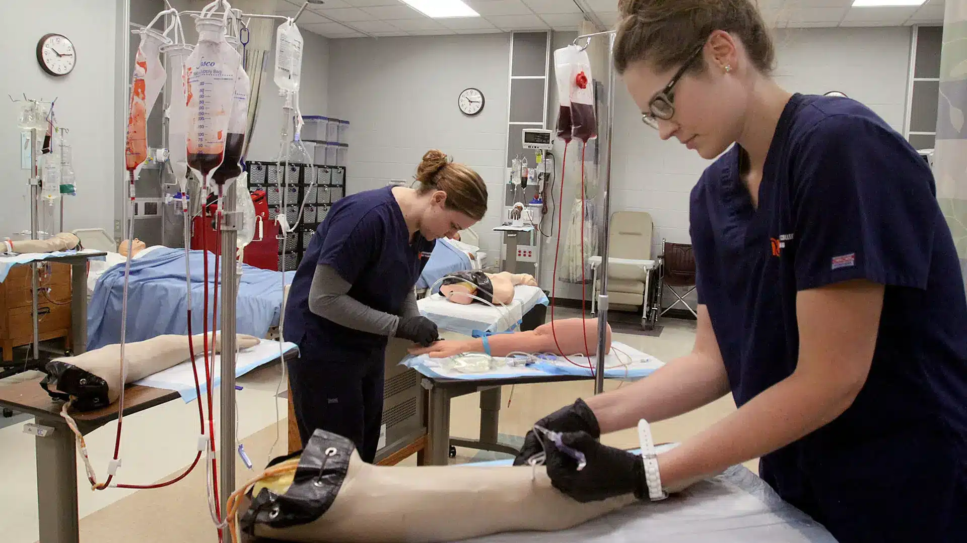 two phlebotomy students practicing taking blood on dummy arms.
