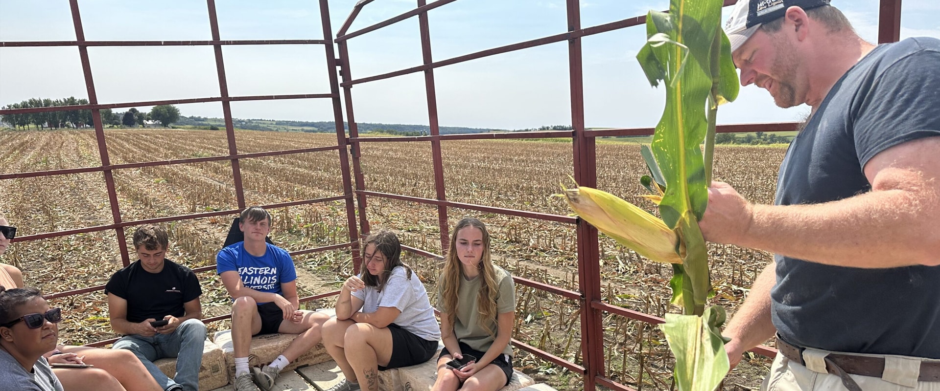 students on a wagon in a field looking at a stalk of corn.